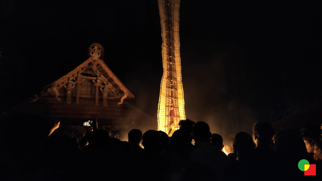A towering lit Theyyam headdress rising over a dark crowd beside the carved gable of a Kerala temple at night.