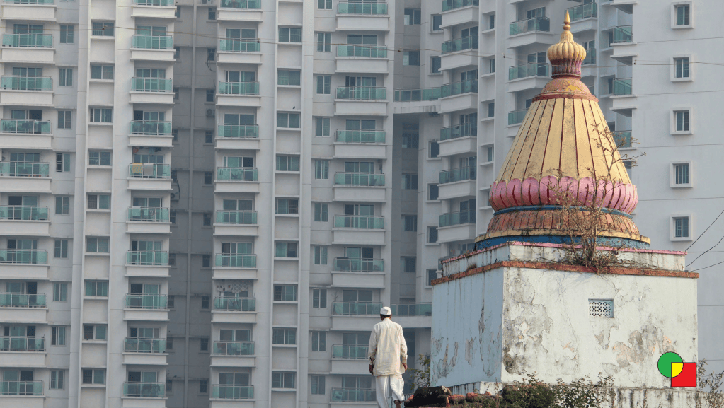 An elderly Maharashtrian man in his traditional attire walks near a small temple in Hinjewadi, with modern high-rise apartment buildings visible in the background.